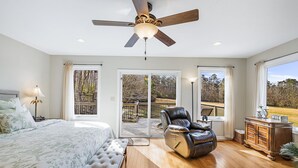 Une chambre à coucher comprenant un lit, un banc, un fauteuil en cuir, une table de chevet, une lampe, un ventilateur de plafond et une vue sur une terrasse accessible par une porte coulissante.