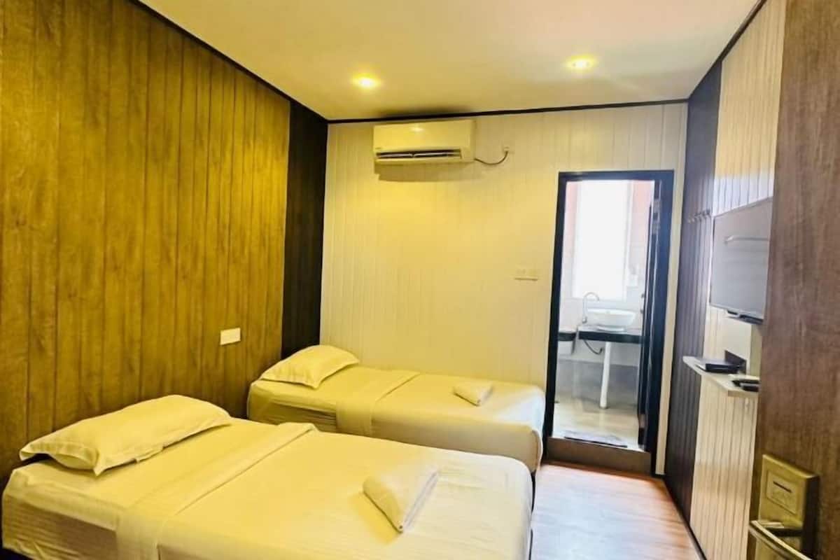 A hotel room with two beds, a wooden wall, an air conditioning unit, and a bathroom visible through a glass door.