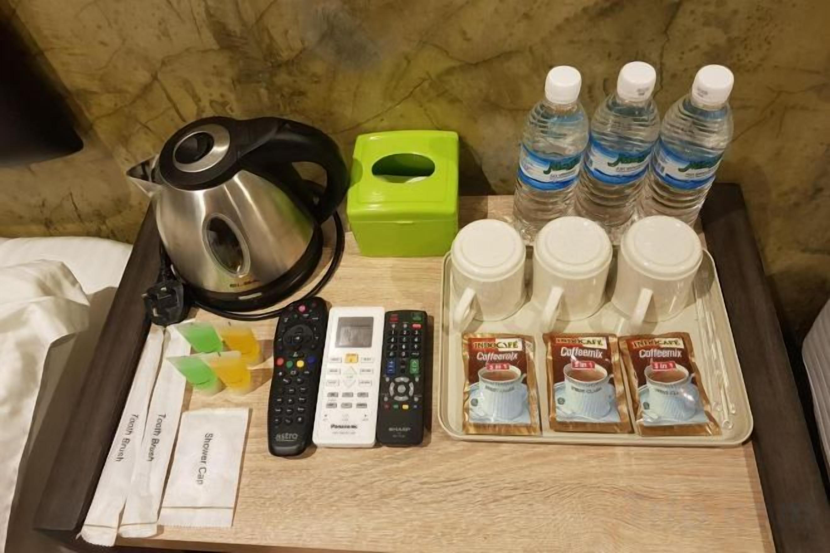 A hotel room's side table with a kettle, remote controls, a green tissue box, bottled water, and coffee packets.