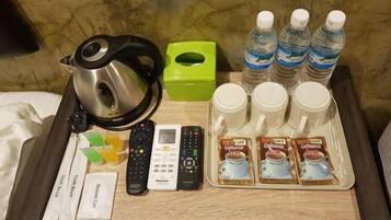 A hotel room's side table with a kettle, remote controls, a green tissue box, bottled water, and coffee packets.