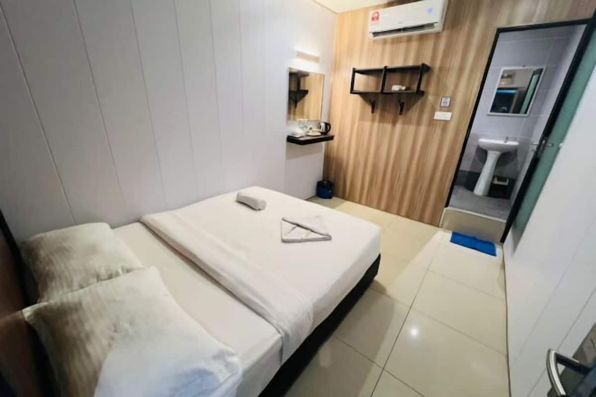 A hotel room with a bed, a small table, a mirror, a shelf with toiletries, and a bathroom visible through an open door.