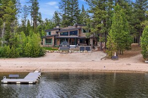 Une maison moderne à deux étages, dotée d’une jetée qui s’avance sur un lac, et entourée d’une forêt dense.