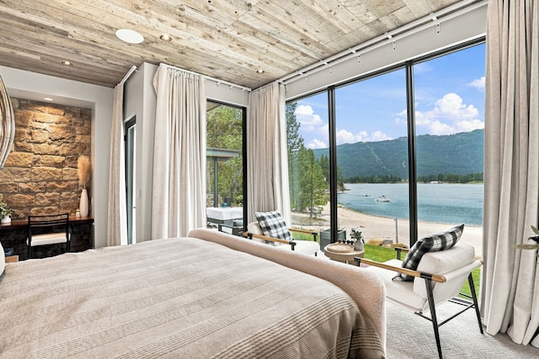 Une chambre avec un grand lit, un mur en pierre, une vue sur un lac et un fauteuil près de la fenêtre.