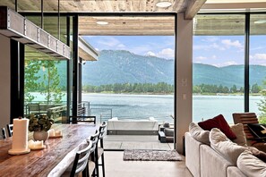 Um interior moderno com área de refeições, um sofá e uma grande janela com vista para um lago e montanhas.