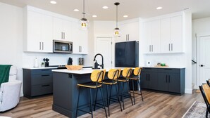 Una cocina moderna con armarios blancos, una isla central con taburetes y electrodomésticos como microondas y refrigerador.
