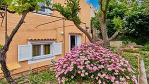 Un edificio con fachada rústica, una ventana con contraventanas y un jardín lleno de flores rosadas.