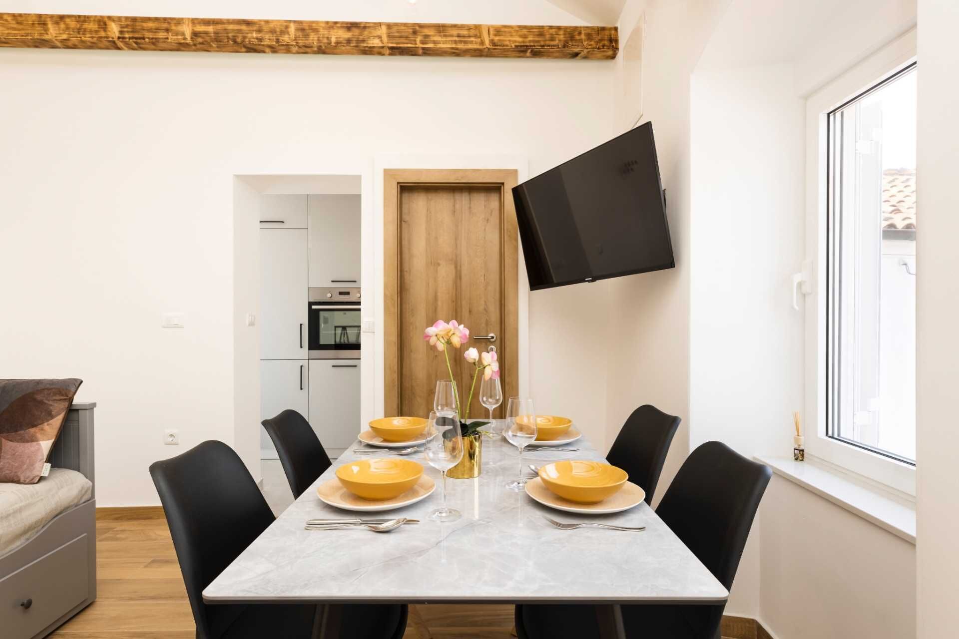 Zona pranzo con tavolo imbandito per sei, sedie, televisore a parete e cucina sullo sfondo.