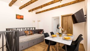 Un espacio habitable moderno con sofá, mesa de comedor y sillas. Cuenta con una zona de cocina con armarios y un televisor montado en la pared.