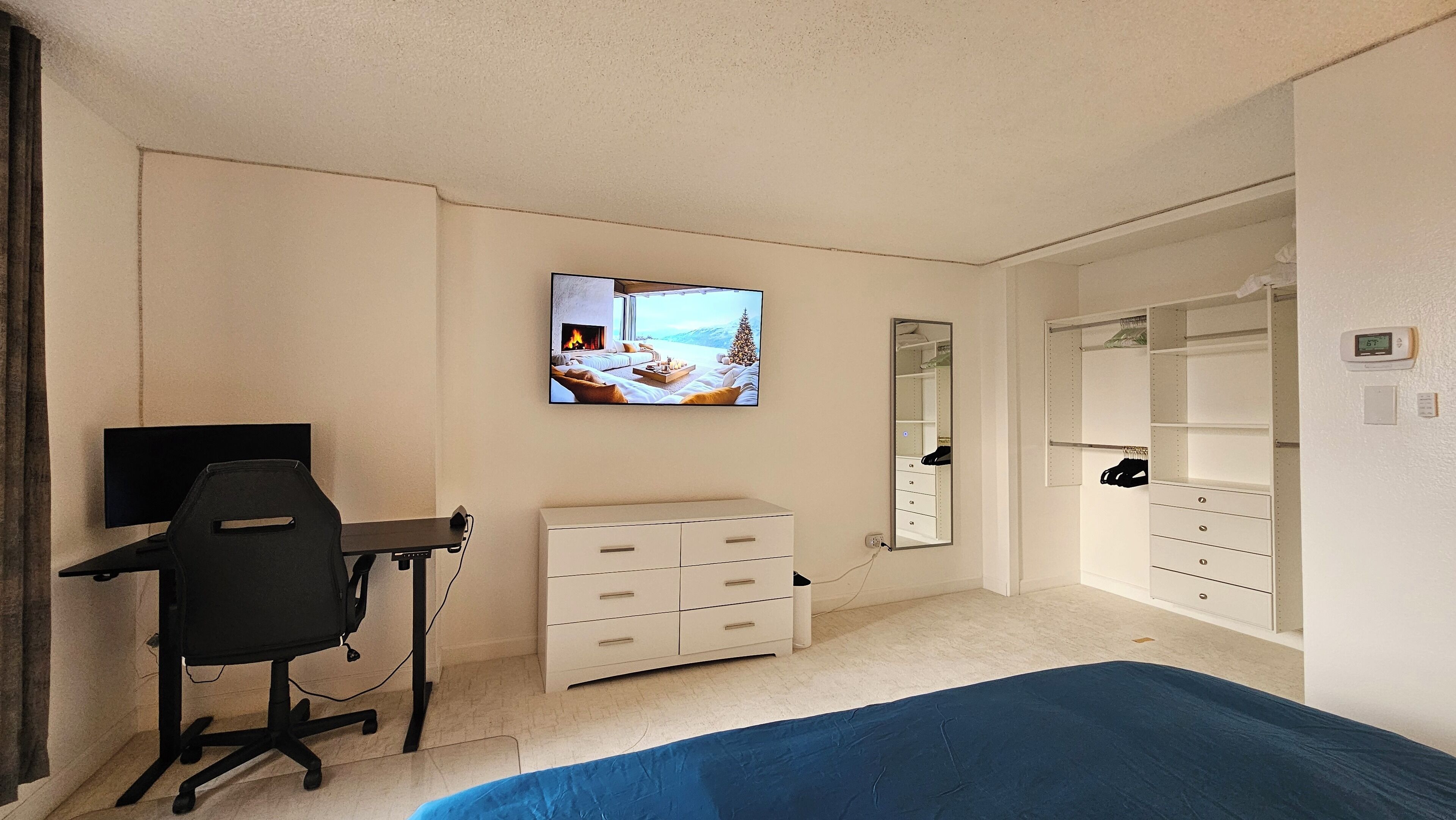Ein Schlafzimmer mit einem Bett, einem Schreibtisch mit Stuhl, einem Fernseher, einer Kommode, einem Kleiderschrank und einem Spiegel.