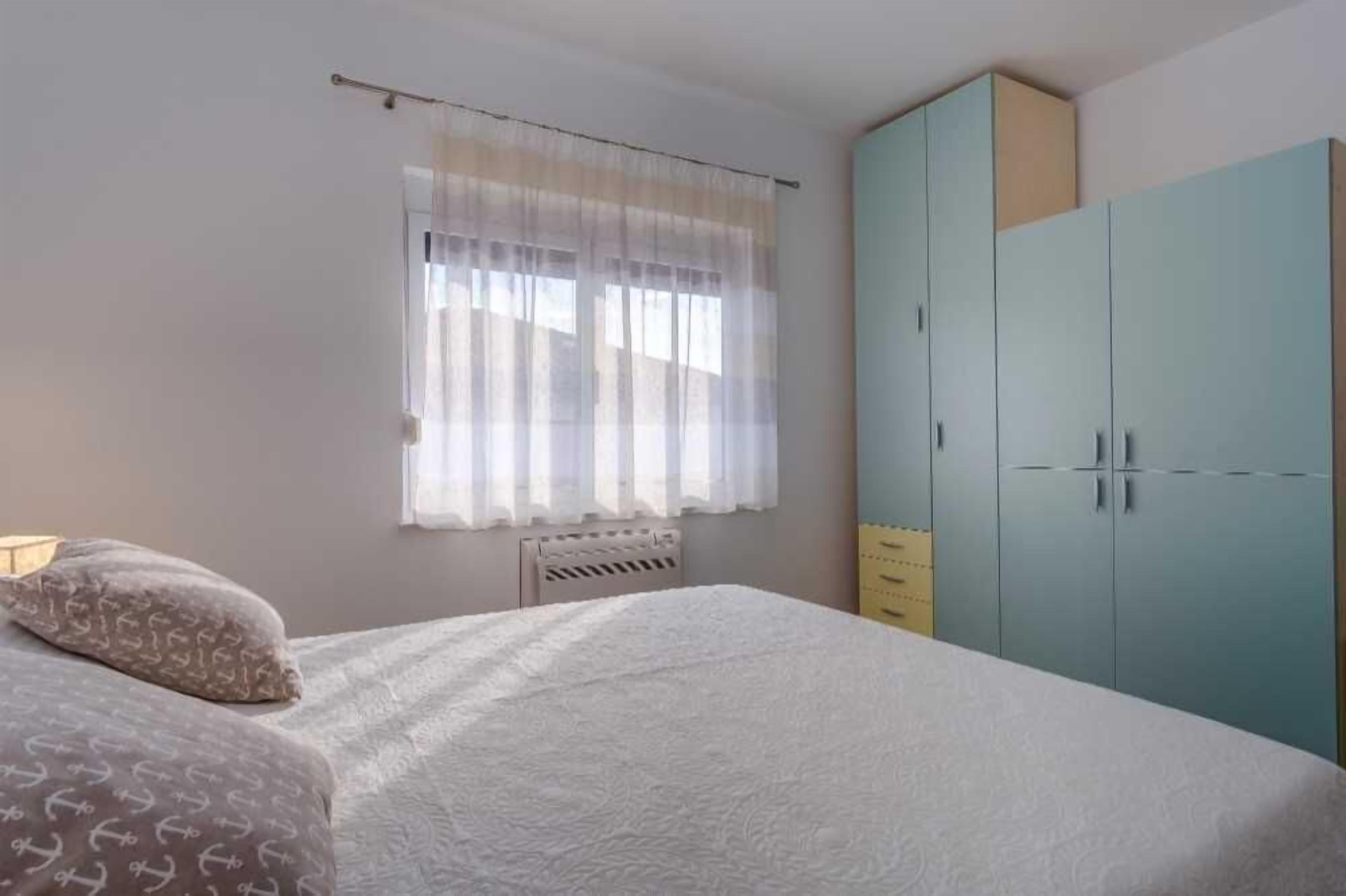 A bedroom with a bed, a window with sheer curtains, a radiator, and a light blue wardrobe.