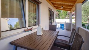 A covered outdoor seating area with a wooden table and wicker chairs, large windows, and a view of a swimming pool. - Cozy 1BR Suite for Three, No Pets, Parking Available (Mali Lošinj)