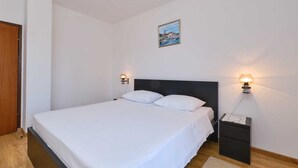En pænt redt seng med hvide sengetæpper i et rum med hvide vægge og trægulv. Der er to vægmonterede lamper på hver side af sengen, og et lille bord med en lampe ved siden af.