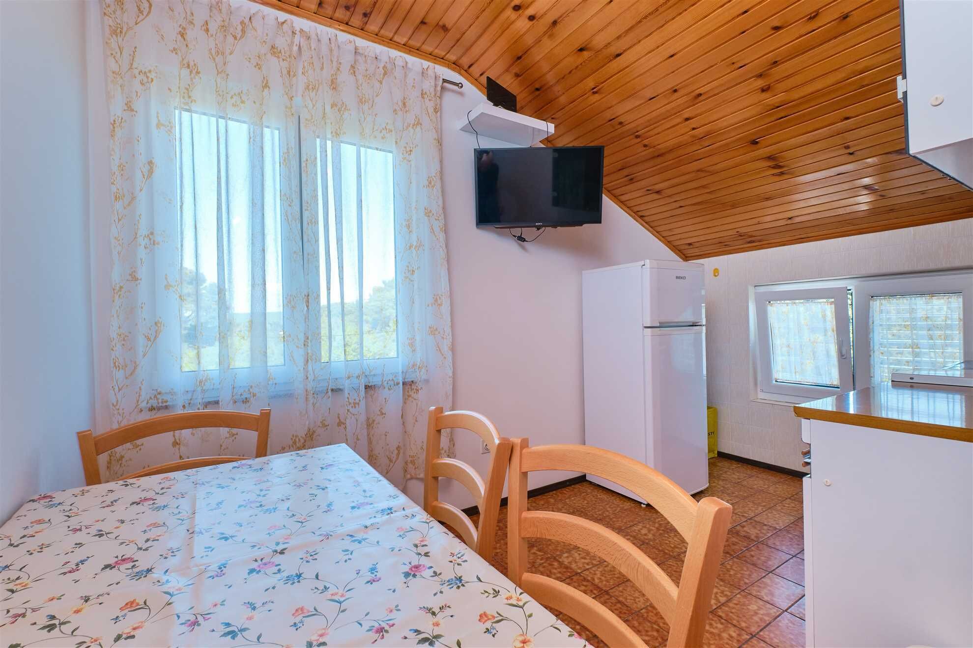 Une pièce avec un plafond en bois, une table à manger recouverte d’une nappe à motifs floraux, deux chaises en bois, un téléviseur fixé au mur, un réfrigérateur et une fenêtre avec des rideaux.