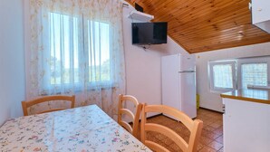 A room with a wooden ceiling, a dining table with floral tablecloth, two wooden chairs, a television mounted on the wall, a refrigerator, and a window with curtains. - Charming 2-Bedrooms Apartment in Nerezine (Nerezine)