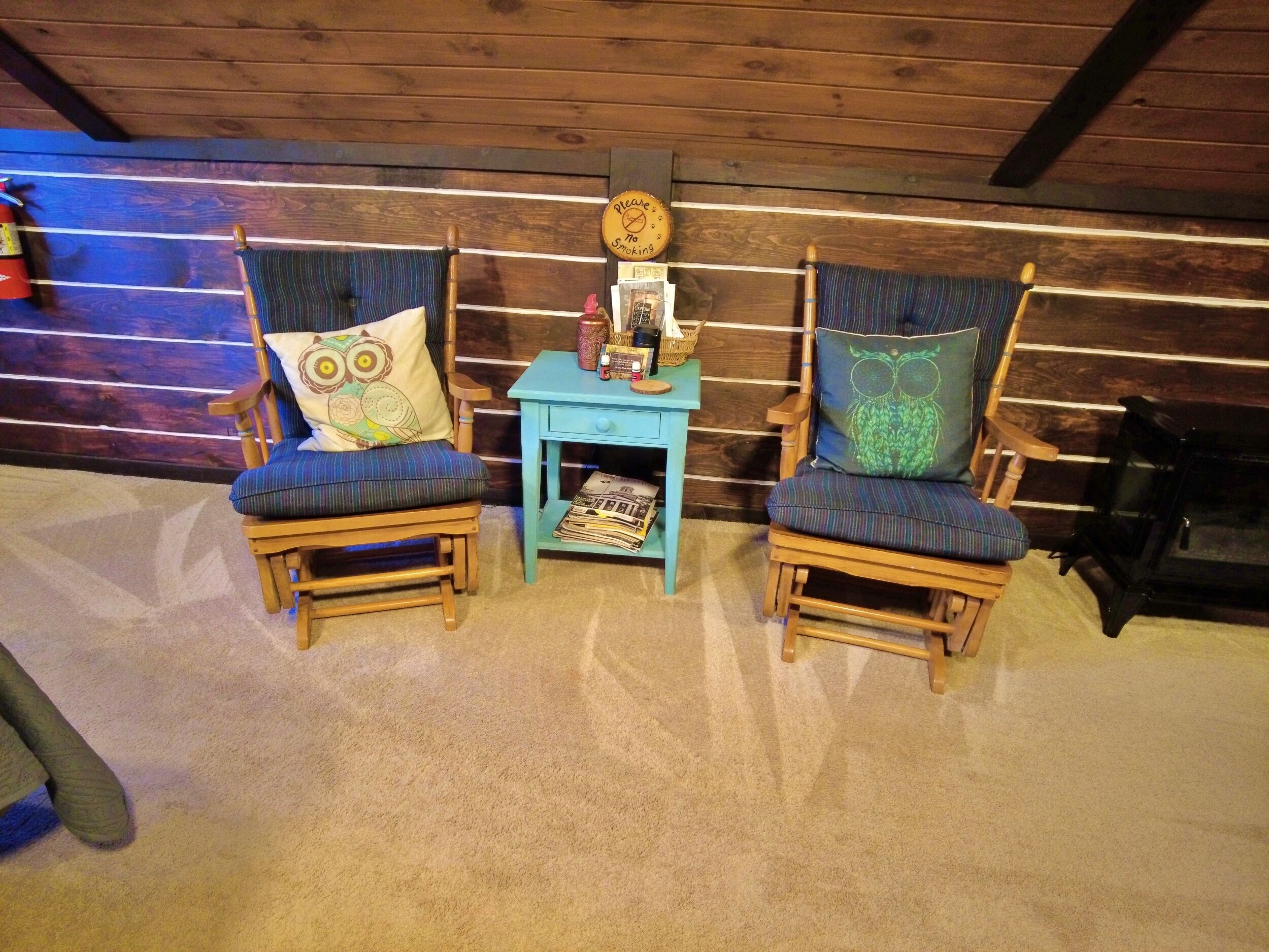 Dos sillas de madera con cojines con motivos de búhos, una mesita auxiliar azul con un reloj y una chimenea en una habitación de estilo cabaña de troncos.