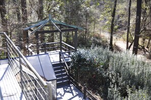 Una terraza de madera con baranda metálica, rodeada de arbustos y árboles frondosos.