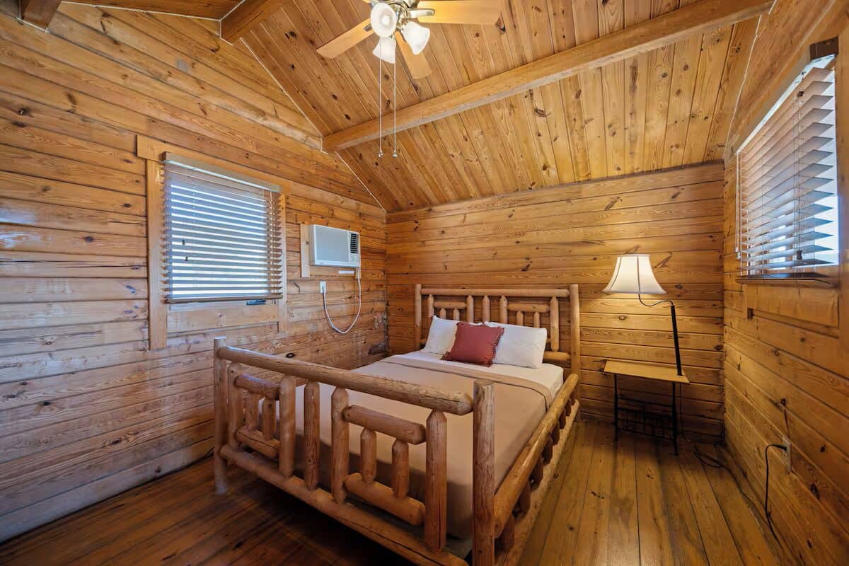 A wooden cabin room with a bed, a ceiling fan, and a window with blinds.