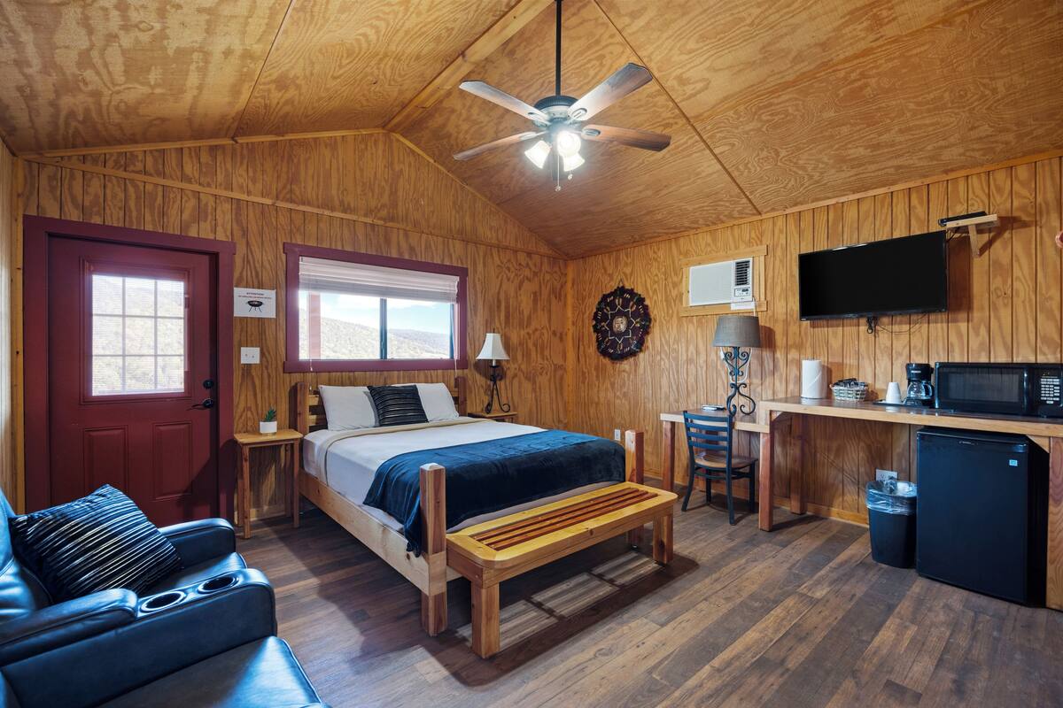 A wooden cabin with a bed, a desk, a television, and a small kitchen area.