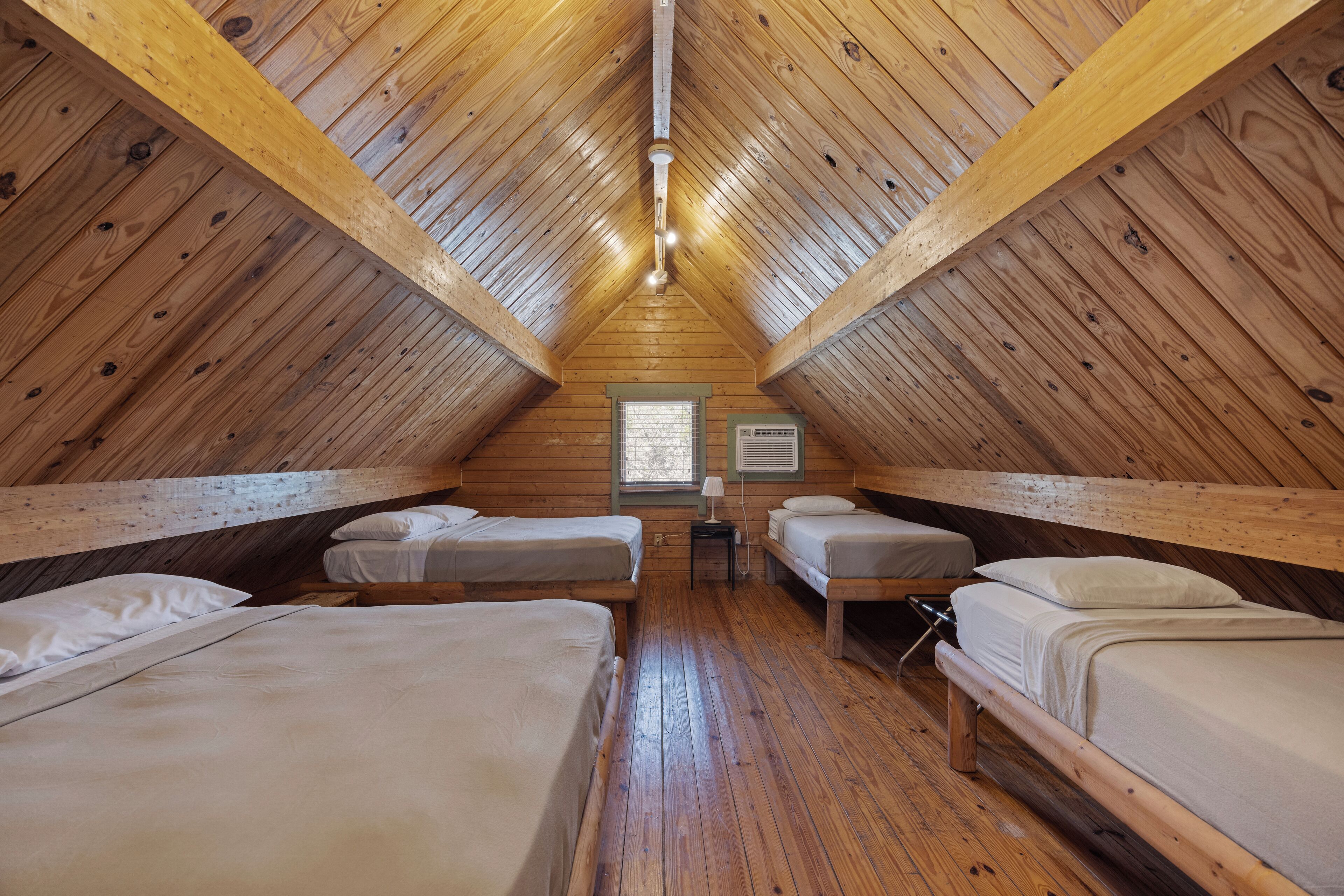 A cozy attic bedroom with four wooden beds arranged in a U-shape, a small table with a lamp, and a window with blinds.