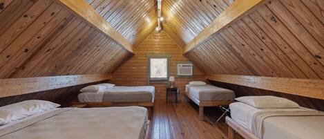 A cozy attic bedroom with four wooden beds arranged in a U-shape, a small table with a lamp, and a window with blinds.
