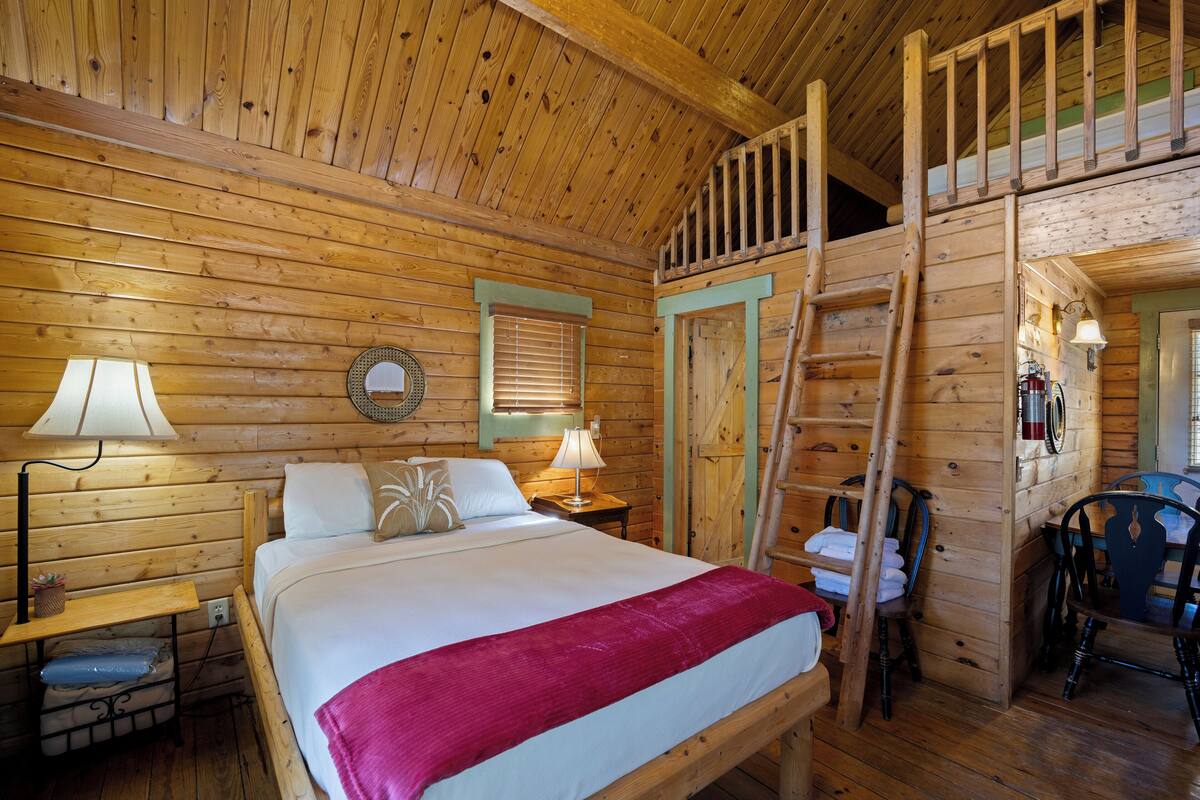 A wooden cabin room with a bed, two bedside tables with lamps, a chair, and a ladder leading to an upper level.