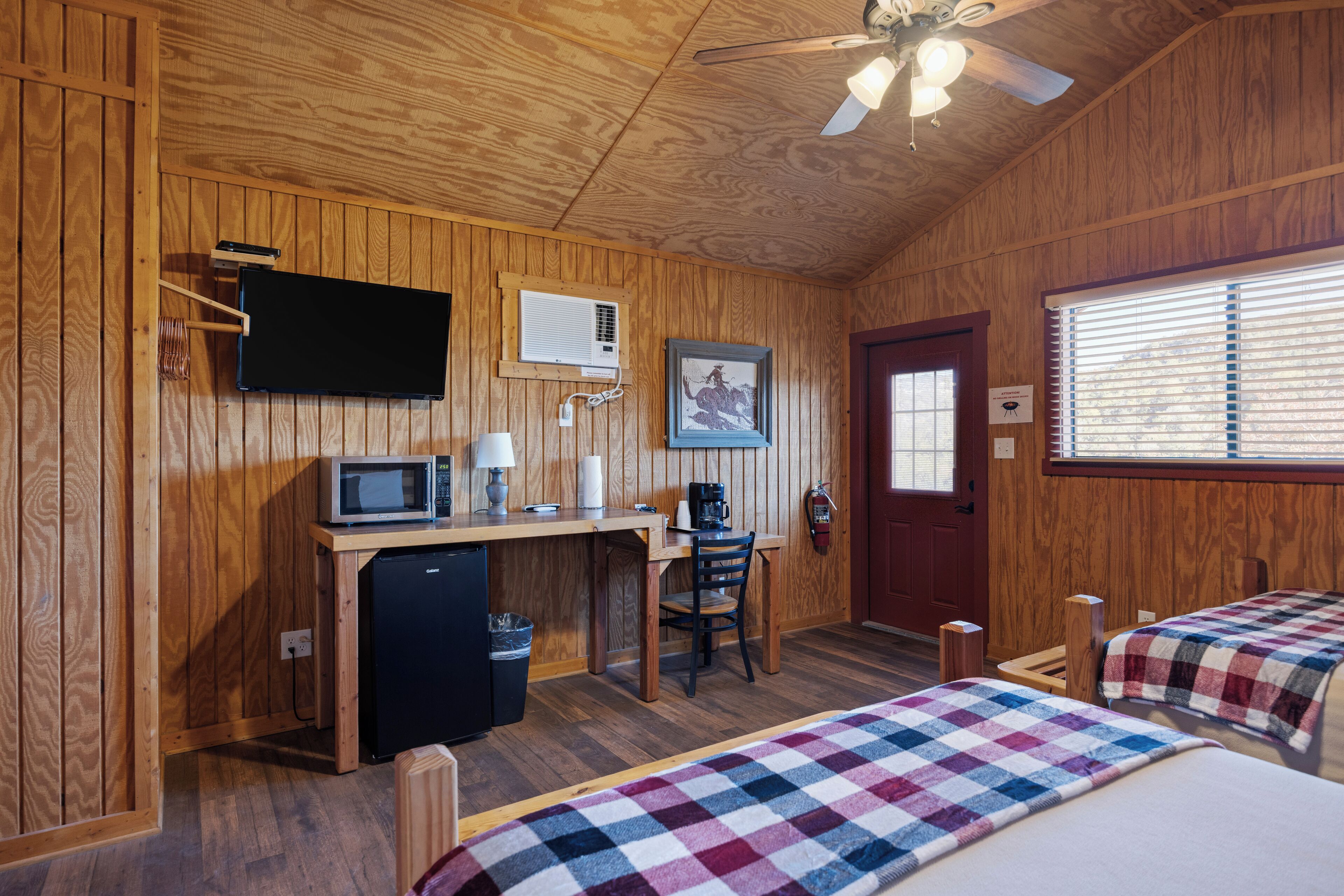 A wooden room with a bed, a desk with a computer, a microwave, and a ceiling fan.