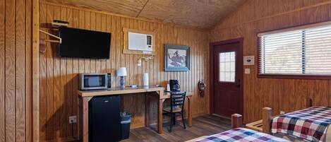 A wooden room with a bed, a desk with a computer, a microwave, and a ceiling fan.