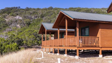 A wooden cabin with a balcony, set in a natural landscape with hills and sparse vegetation.