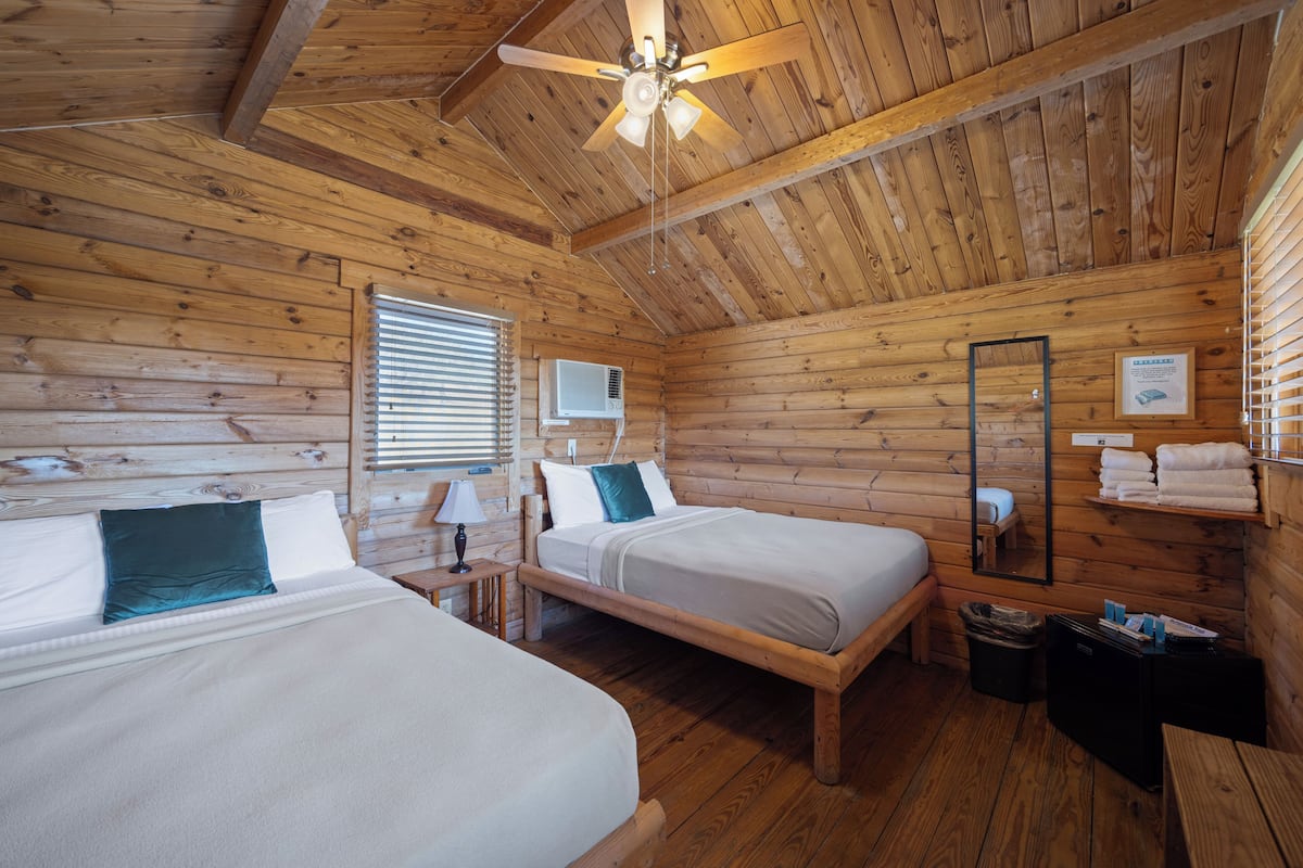 A wooden cabin room with two beds, a ceiling fan, a window with blinds, an air conditioning unit, and a mirror on the wall.
