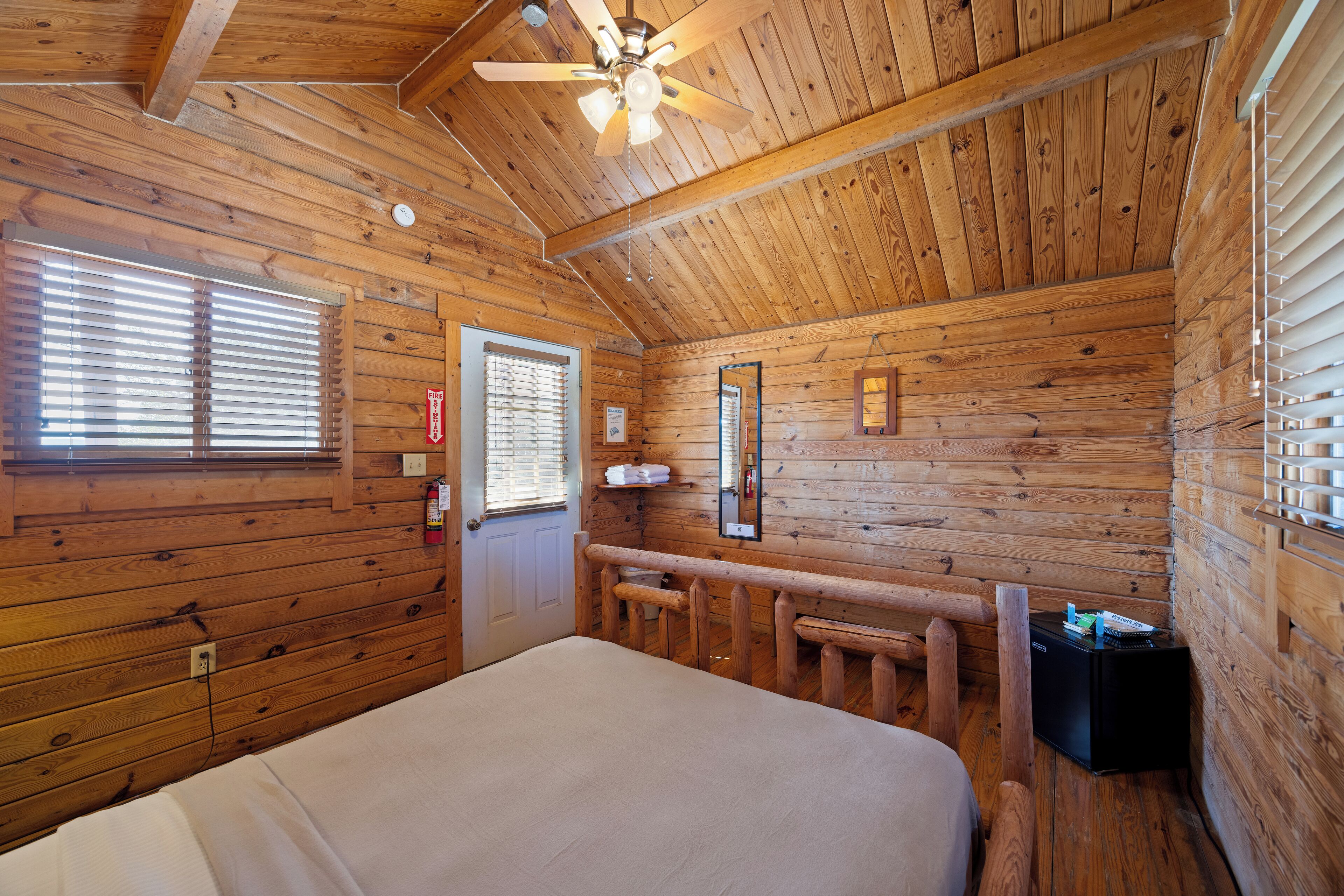 A wooden cabin room with a bed, a ceiling fan, a fire extinguisher, and a window with blinds.