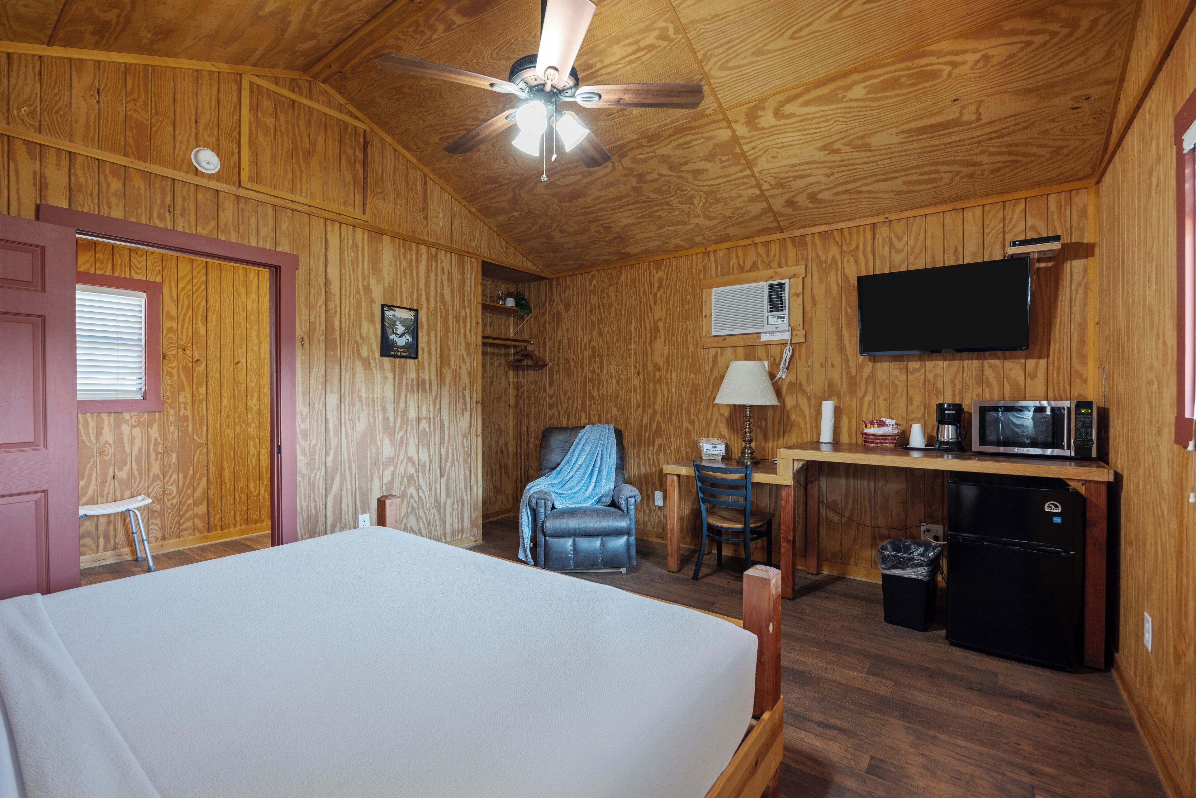 Een kamer met houten lambrisering, een bed, een bureau met een computer, een stoel, een plafondventilator en een televisie.