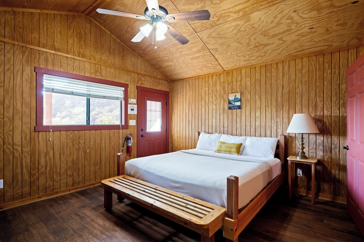 A wooden-paneled bedroom with a bed, bench, bedside table, lamp, and a ceiling fan.