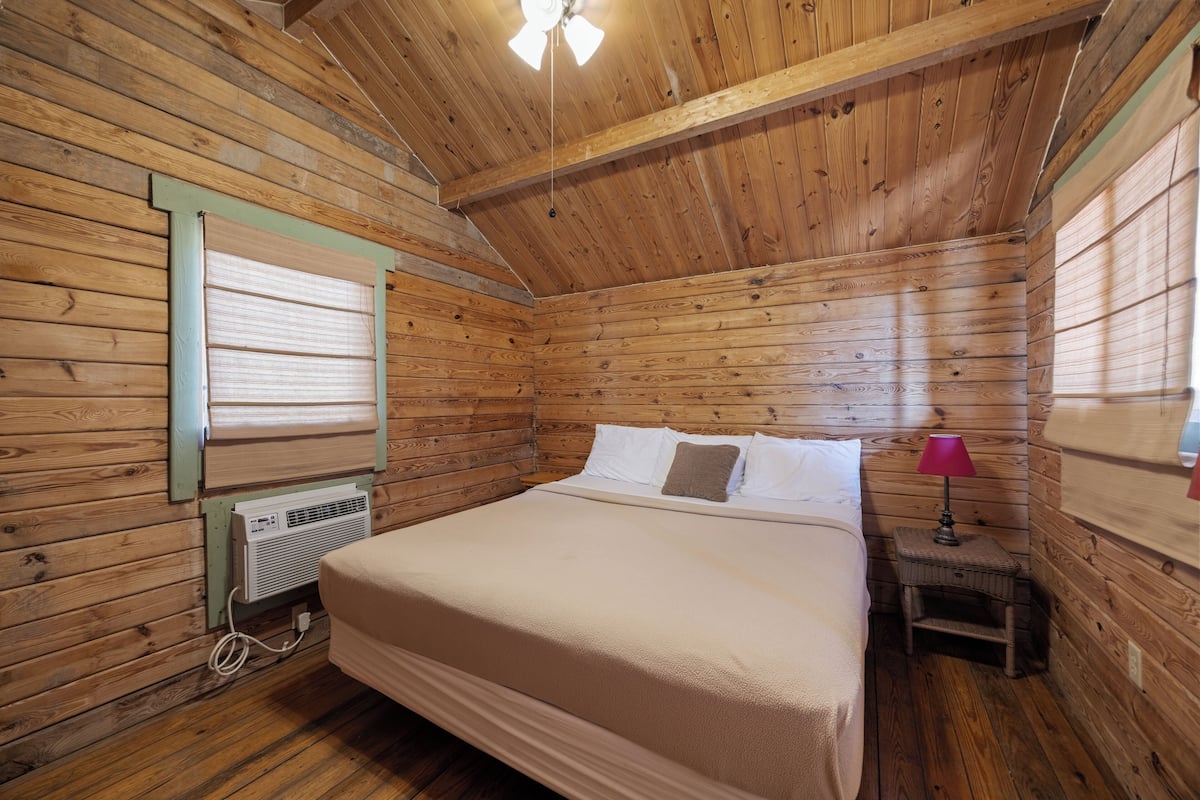 A wooden cabin bedroom with a bed, a nightstand, a lamp, and a window with blinds.
