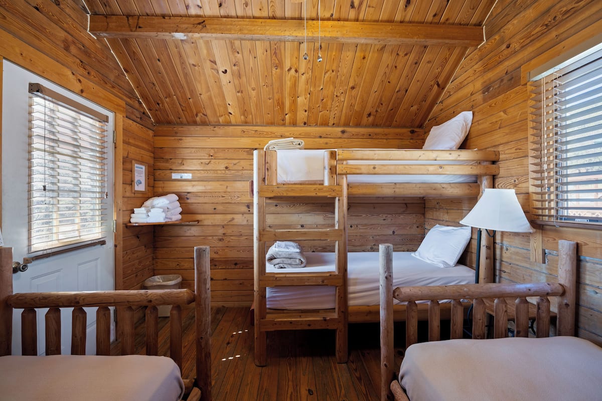 A wooden cabin with bunk beds, a small shelf with towels, and a window with blinds.