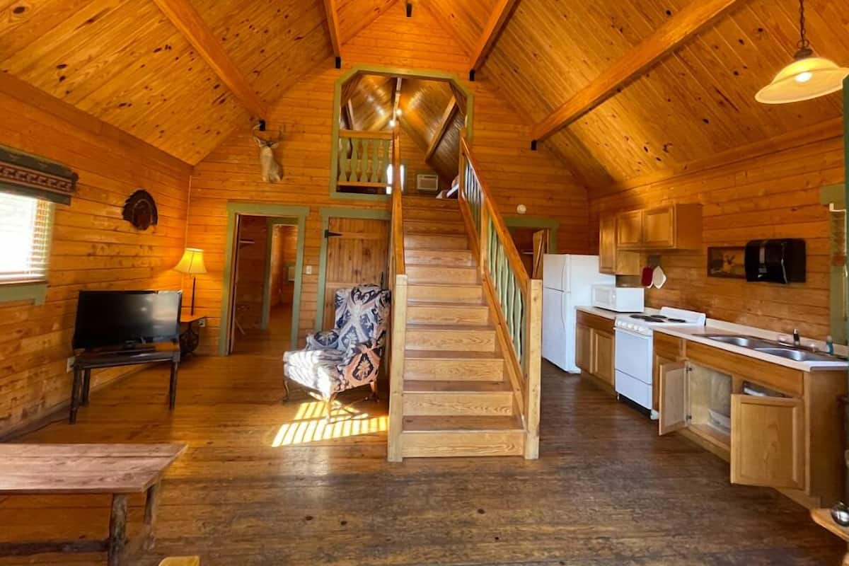 A wooden cabin interior with a staircase leading to a loft bedroom, a kitchen area, and a seating area with a bench and a lamp.