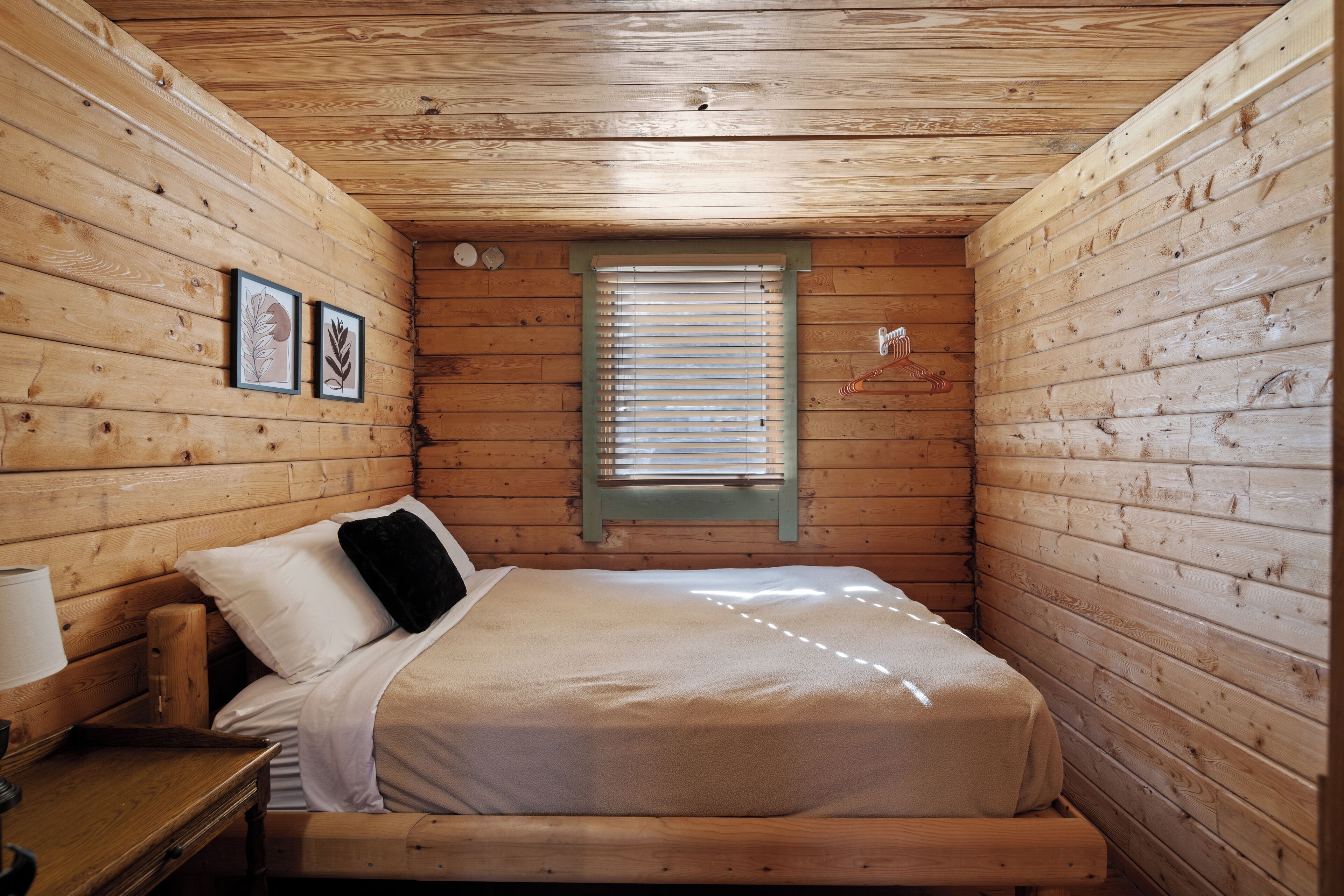 Ein Zimmer in einer Holzhütte mit einem Bett, einem Nachttisch, einem Fenster mit Jalousien und zwei gerahmten Bildern an der Wand.