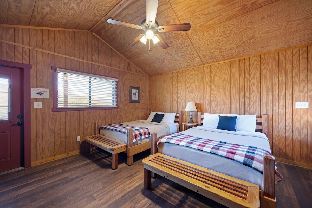 A wooden-paneled room with two beds, a ceiling fan, and a window with blinds.