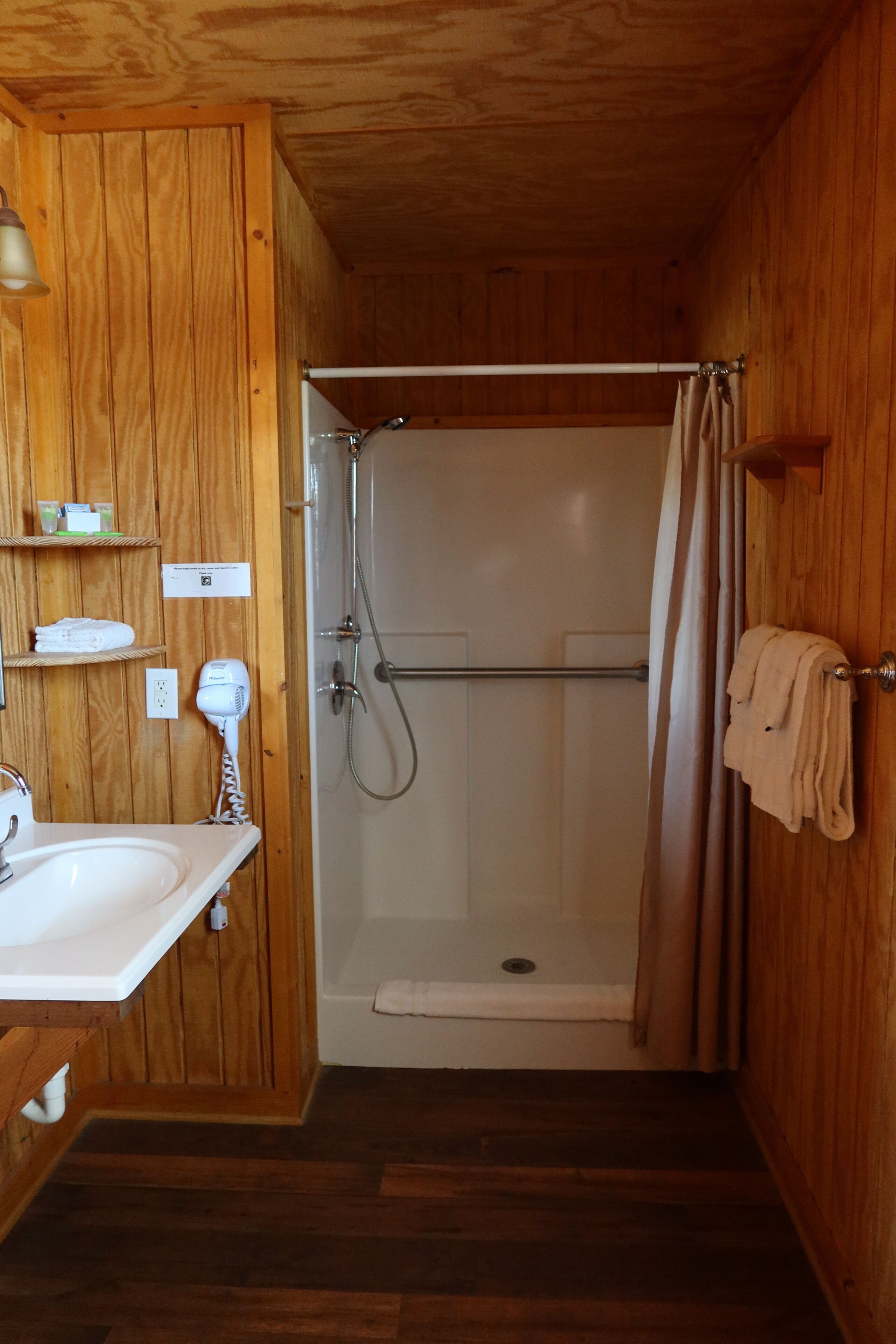 A wooden-paneled bathroom with a sink, a hairdryer, a shower with a curtain, and a towel rack.