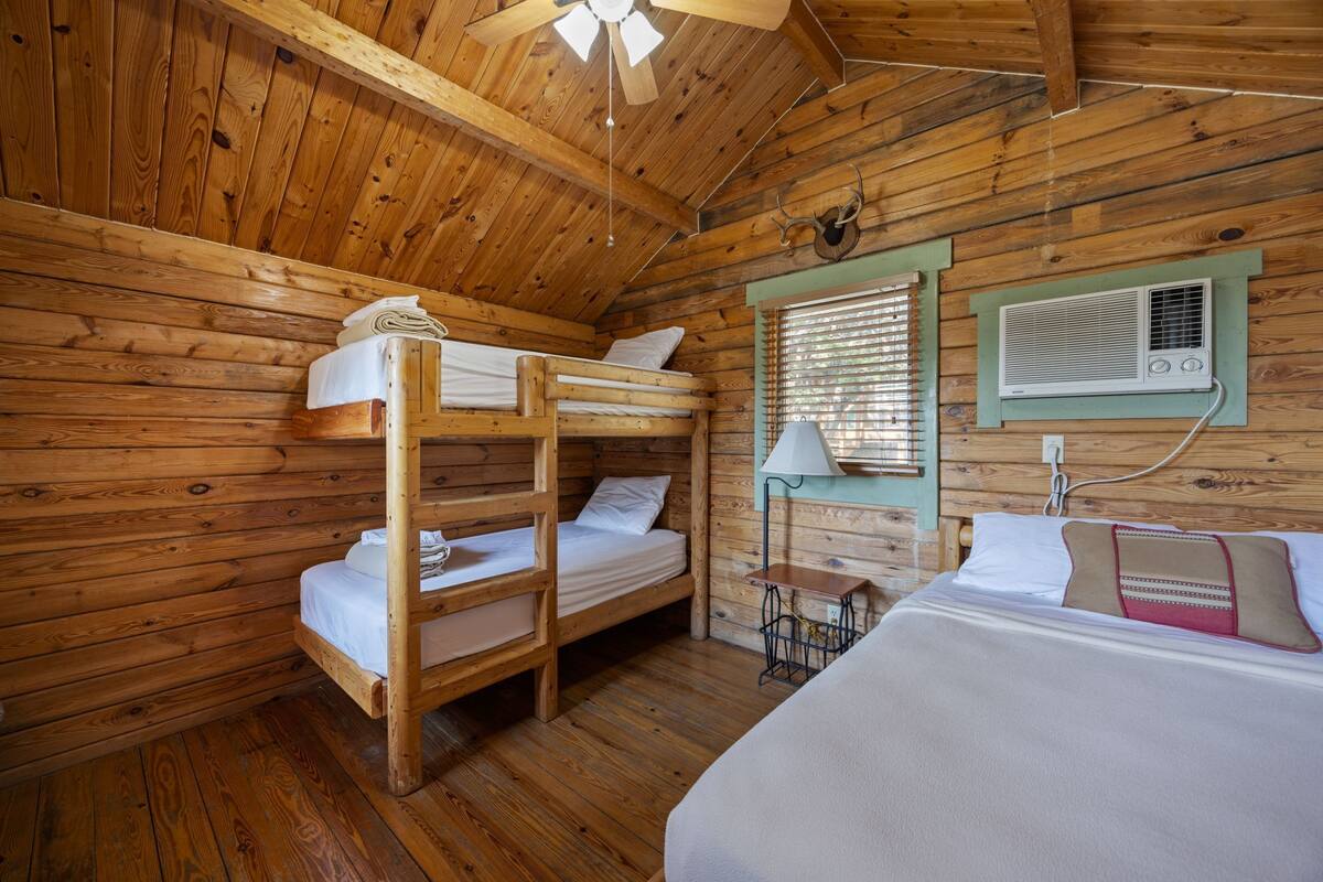 A wooden cabin room with bunk beds, a single bed, a window with blinds, a wall-mounted air conditioner, and a small table with a lamp.