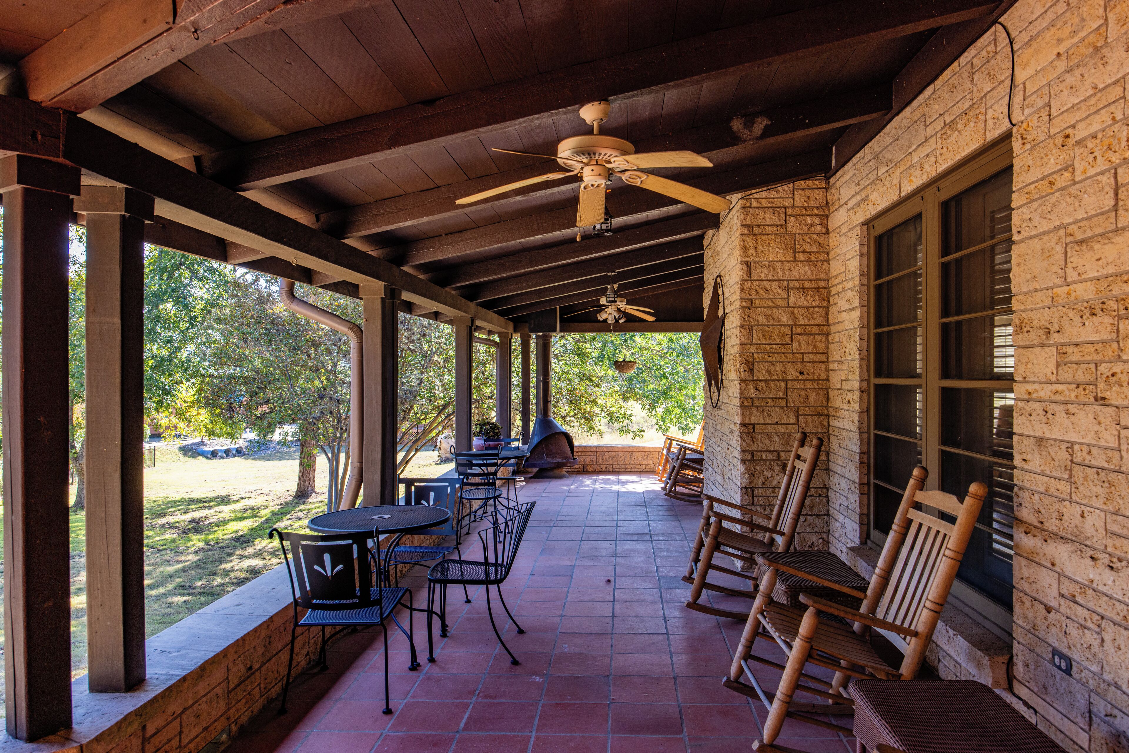 A covered patio with wooden furniture, a ceiling fan, and a brick wall.