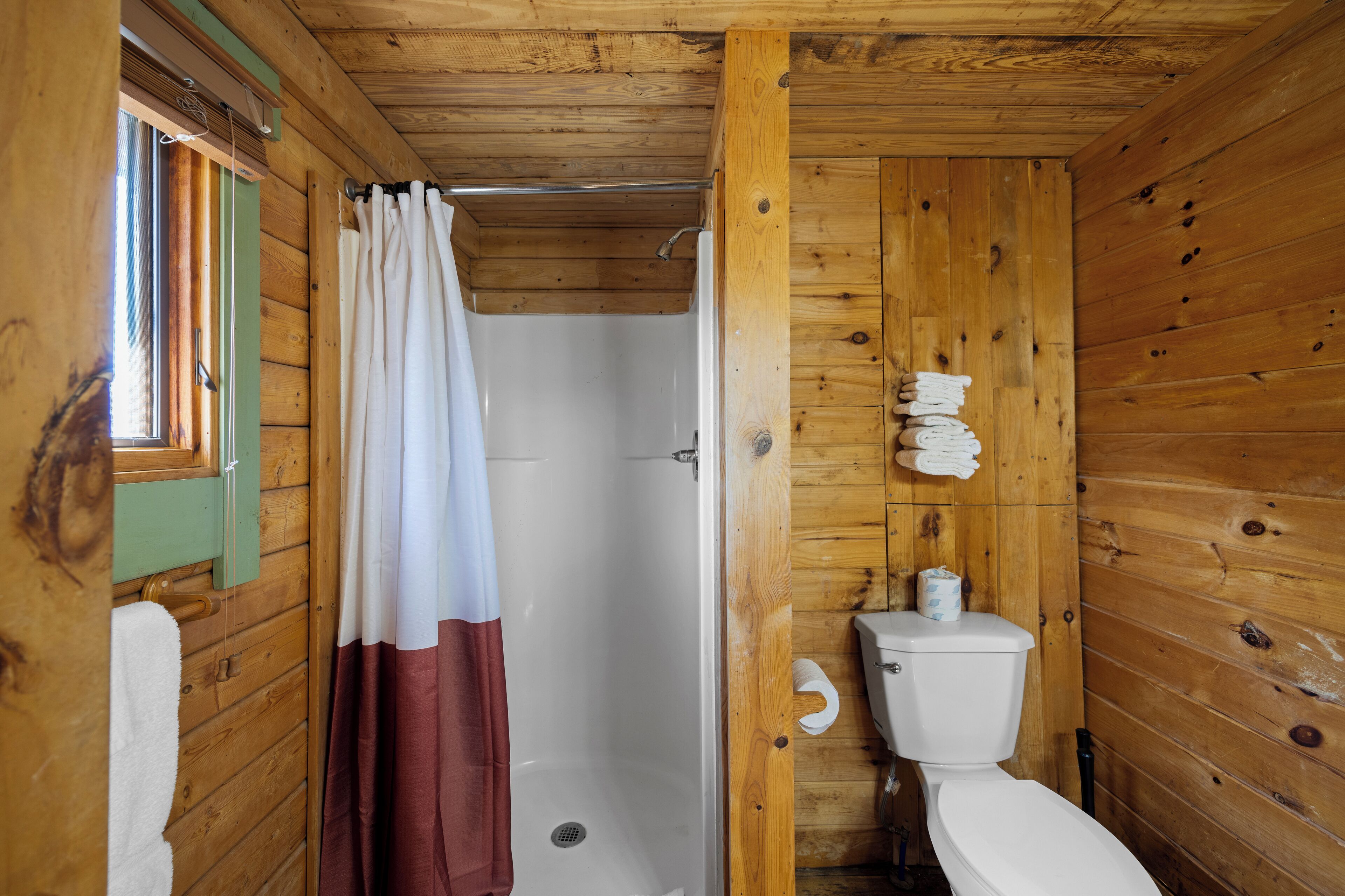 Baño de cabaña de madera con ducha, inodoro y ventana con cortinas.