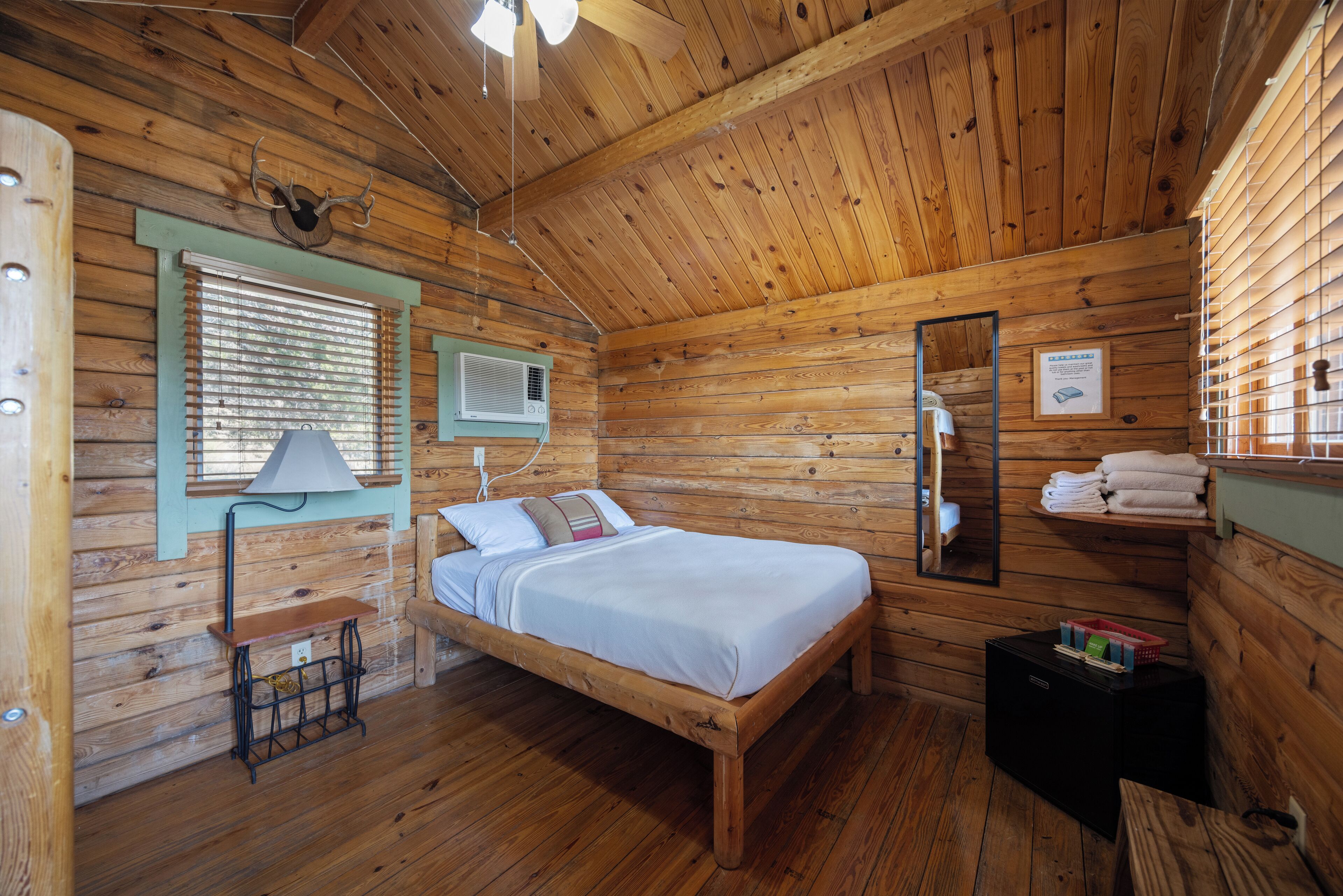 Une chambre dans un chalet en bois, comprenant un lit, une fenêtre avec des stores, un appareil de climatisation, un miroir et une étagère avec des serviettes.