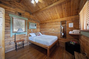 A wooden cabin room with a bed, a window with blinds, an air conditioning unit, a mirror, and a shelf with towels. - Texas Hill Country Resort (Medina)