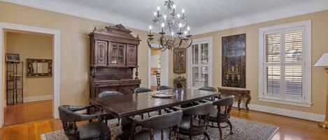A dining room with a large wooden table, six chairs, a chandelier, and a vintage piano.