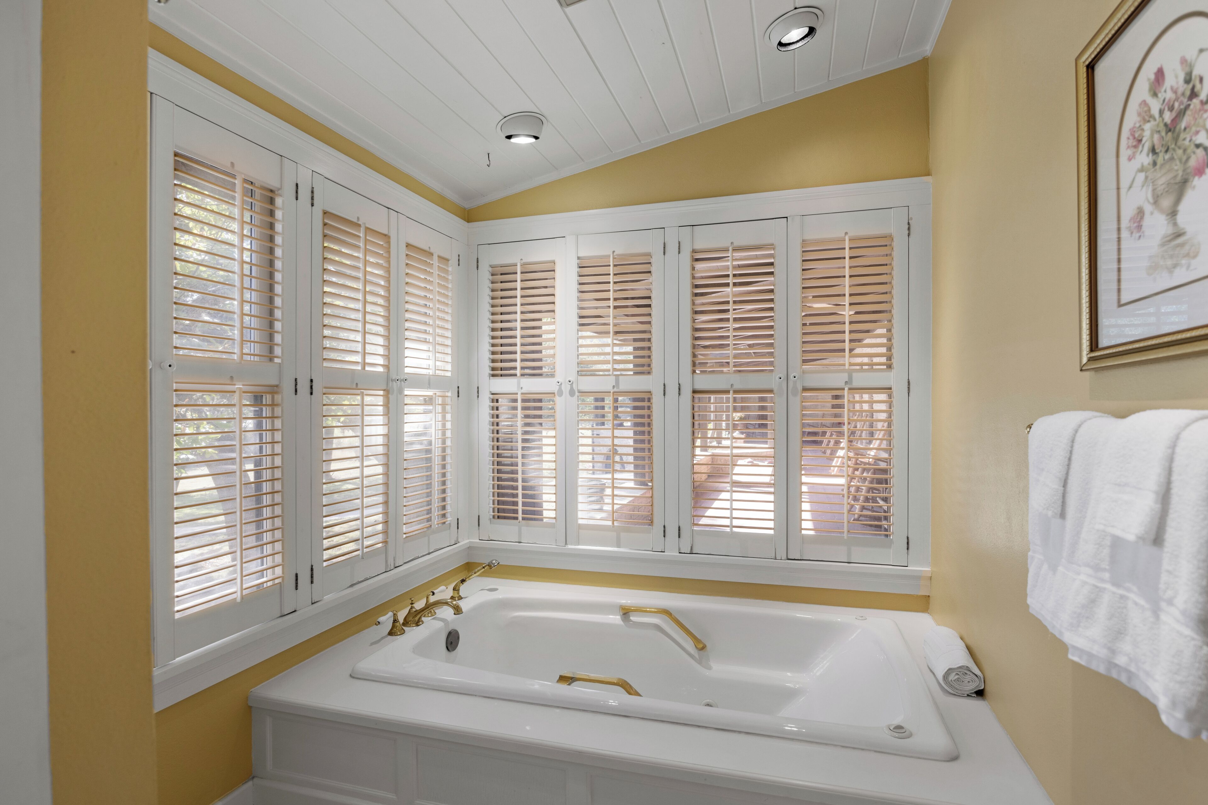 Une salle de bain avec une baignoire blanche, des volets en bois aux fenêtres et un tableau encadré au mur.