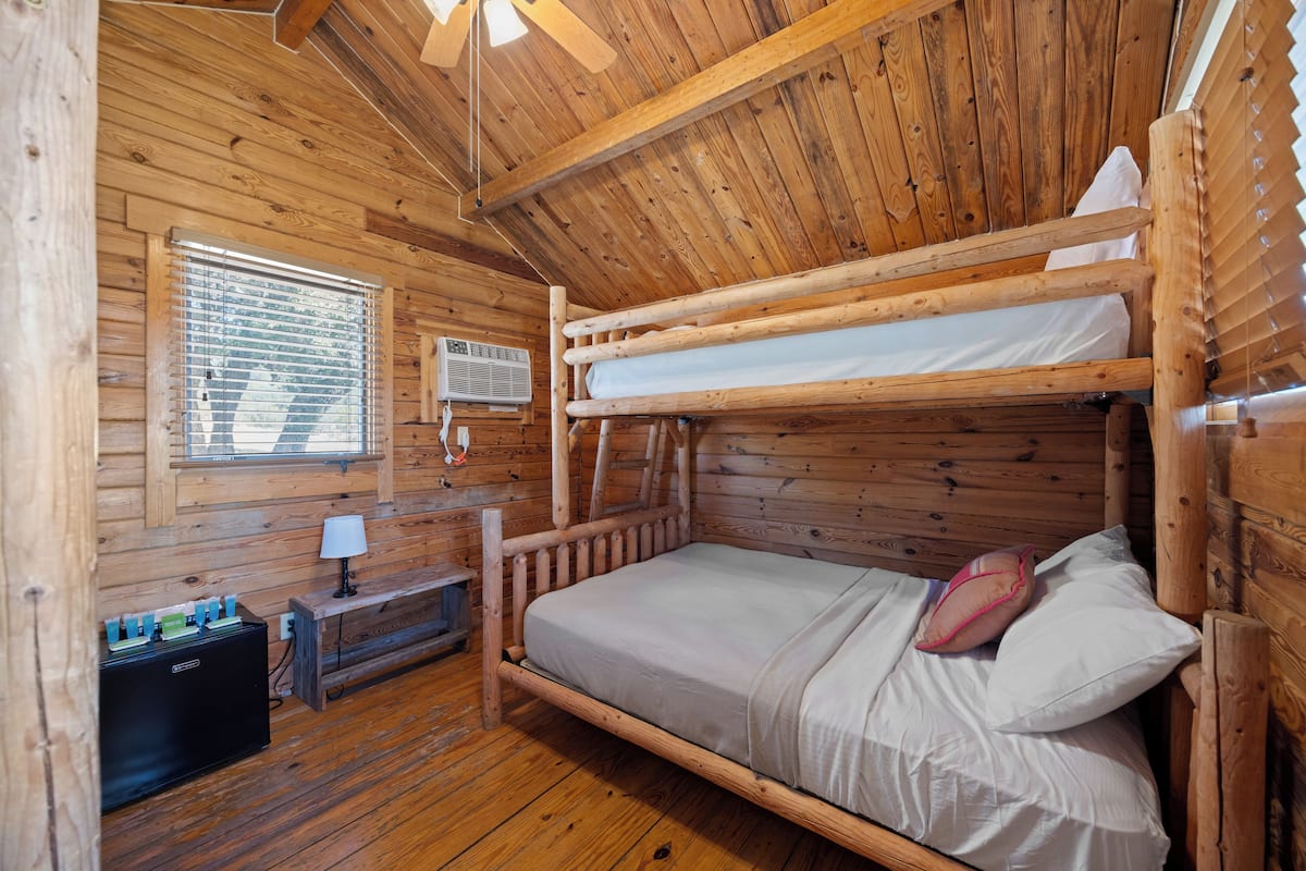 A wooden cabin room with a bunk bed, a small table with a lamp, and a heater on the floor.