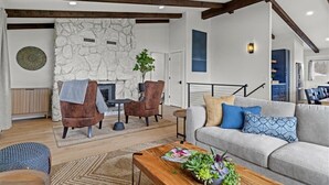Una sala de estar moderna con chimenea de piedra, vigas de madera, un sofá gris y una mesa de centro con una revista y plantas.