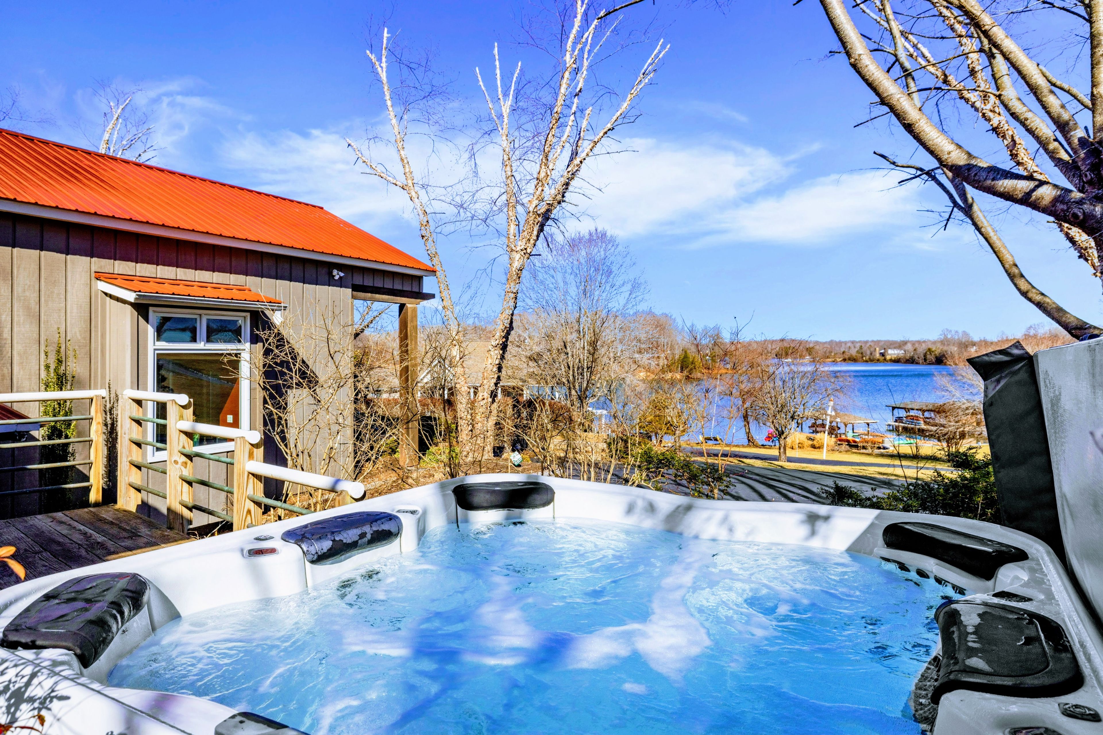 Un bain à remous avec une terrasse en bois, un bâtiment en bois doté d’un toit rouge et un lac en arrière-plan.