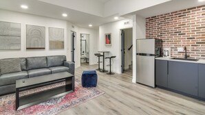 Un salon moderne avec un canapé gris, une table basse et un tapis à motifs. On y trouve également un coin cuisine avec un mur de fond en briques et un réfrigérateur.