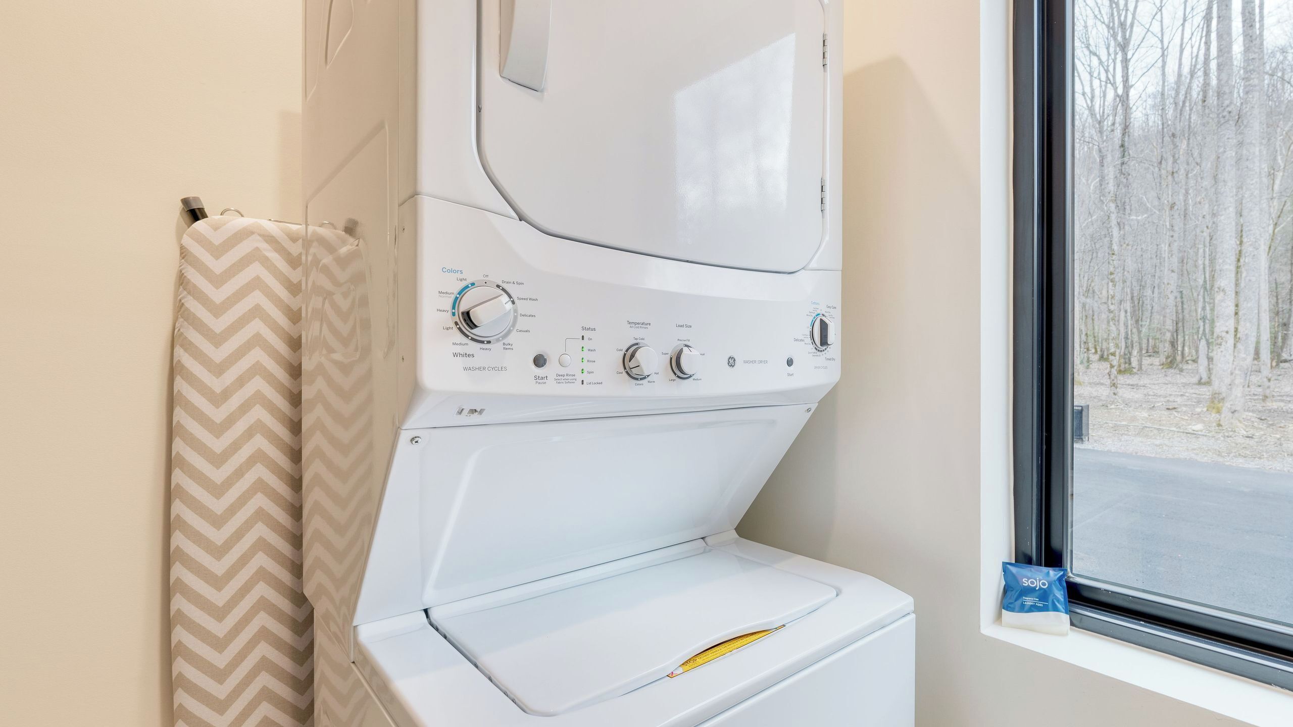 Un appareil compact combinant lave-linge et sèche-linge, doté de boutons de commande, d’une fenêtre donnant sur les arbres et d’une serviette à motifs chevrons.